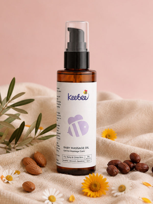 A bottle of Keebee Baby Massage Oil in an amber glass bottle with a black pump and clear cap, set on a beige fabric with almonds, daisies, and yellow flowers against a soft pink background.